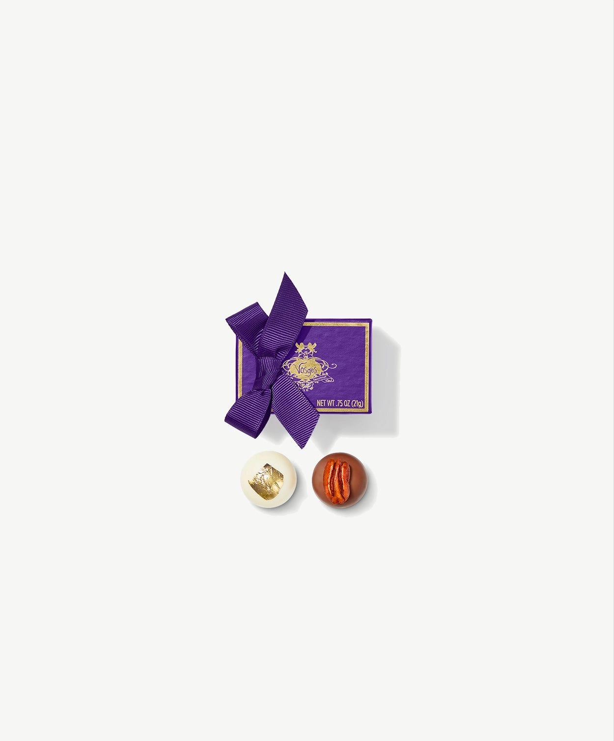 A Vosges Ambrosia truffle and Wink of the Rabbit truffle adorned in gold leaf and a mexican pecan sit side by side in-front a small purple candy box tied with a purple ribbon bow on a grey background.