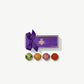 Four Vosges Exotic chocolate truffles dusted with orange curry powder and green matcha sit infront a rectangular purple candy box embossed with gold foil tied with a purple ribbon bow on a grey background.