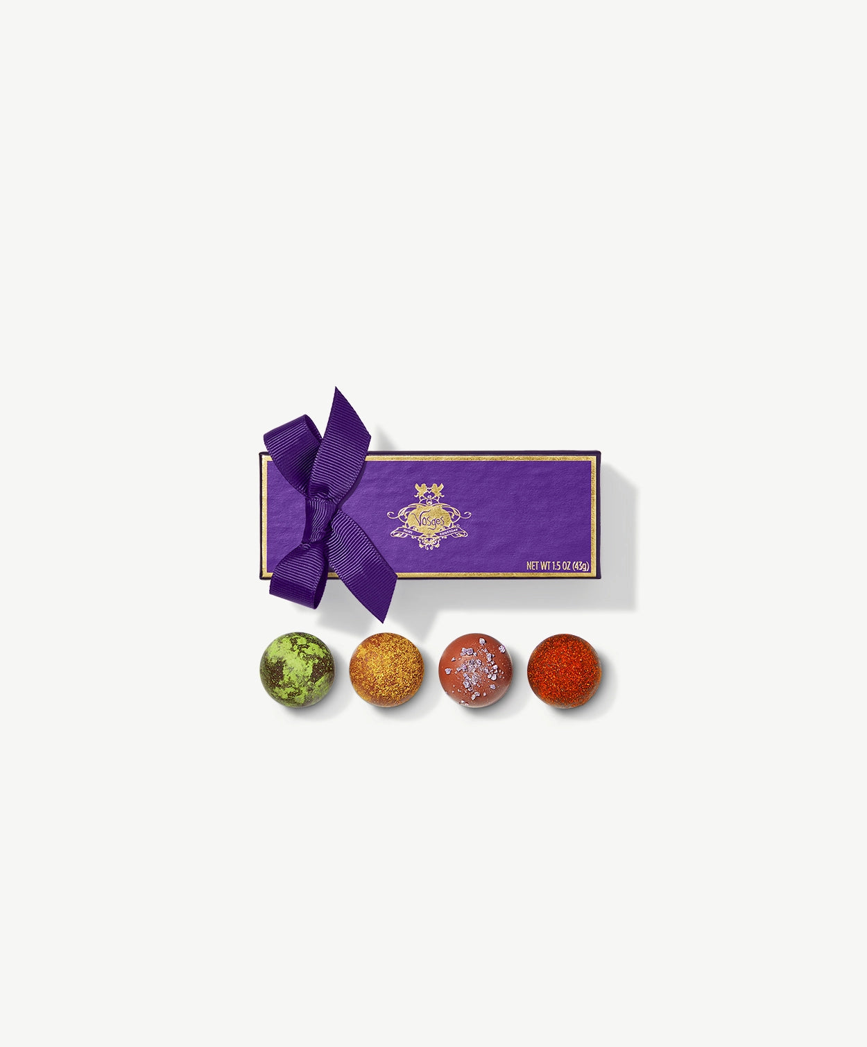 Four Vosges Exotic chocolate truffles dusted with orange curry powder and green matcha sit infront a rectangular purple candy box embossed with gold foil tied with a purple ribbon bow on a grey background.