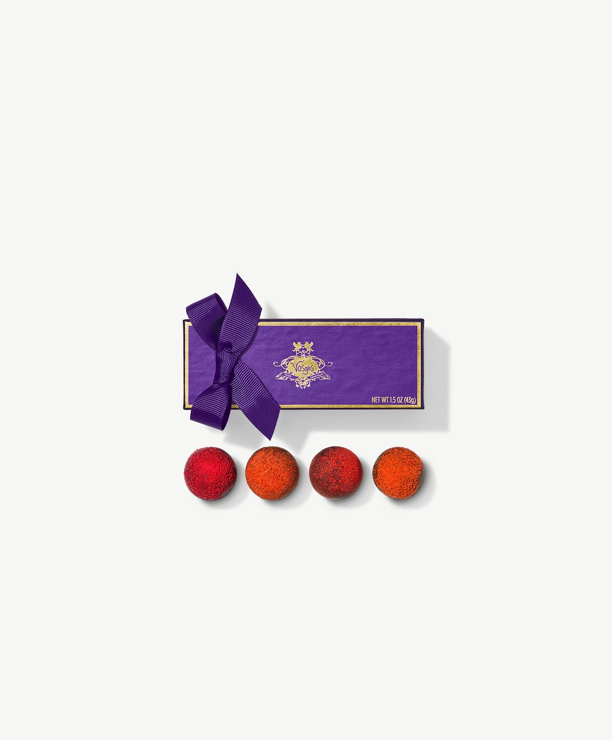 Four Vosges Vegan chocolate truffles adorned with vibrant red raspberry powder and candied orange peel sit infront a rectangular purple candy box embossed with gold foil tied with a purple ribbon bow on a grey background.
