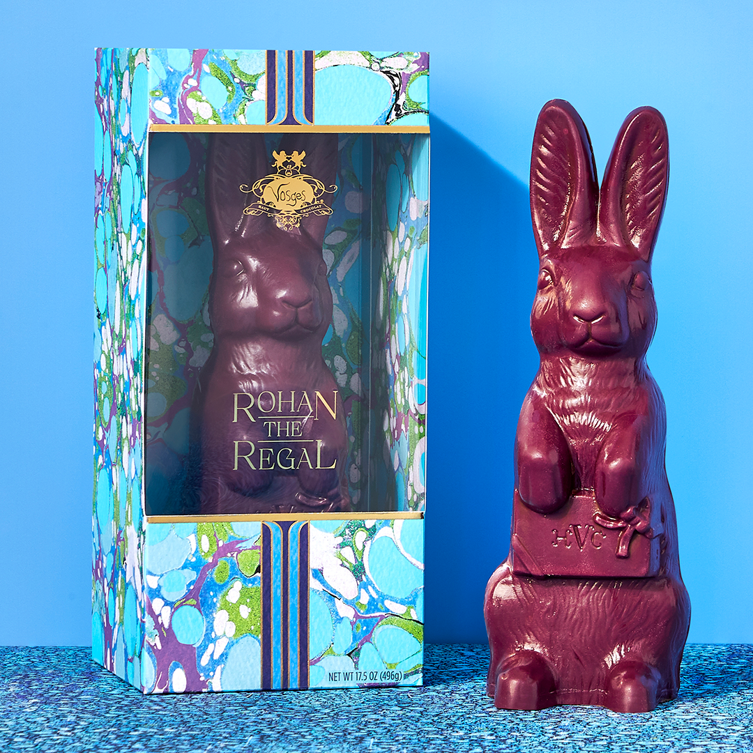 Two identical large purple chocolate rabbits stand side by side, one in a sky blue candy box, on a bright blue background.