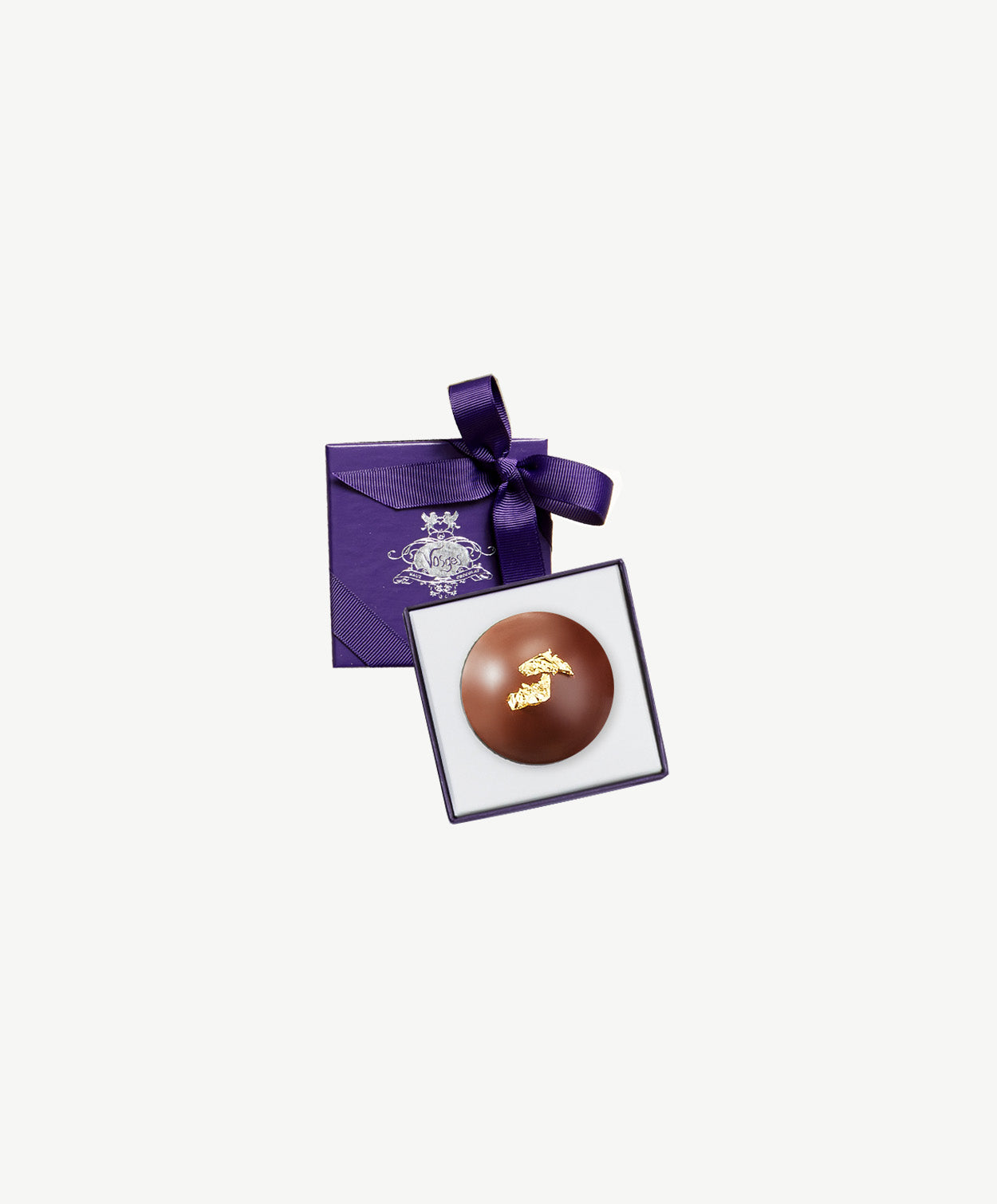 A large hazelnut chocolate bombalina truffle made with a blend of chaga, lion's mane, cordyceps, reishi, maitake, turkey tail mushrooms, sits in a small white and purple candy box tied with a purple ribbon bow on a grey background.