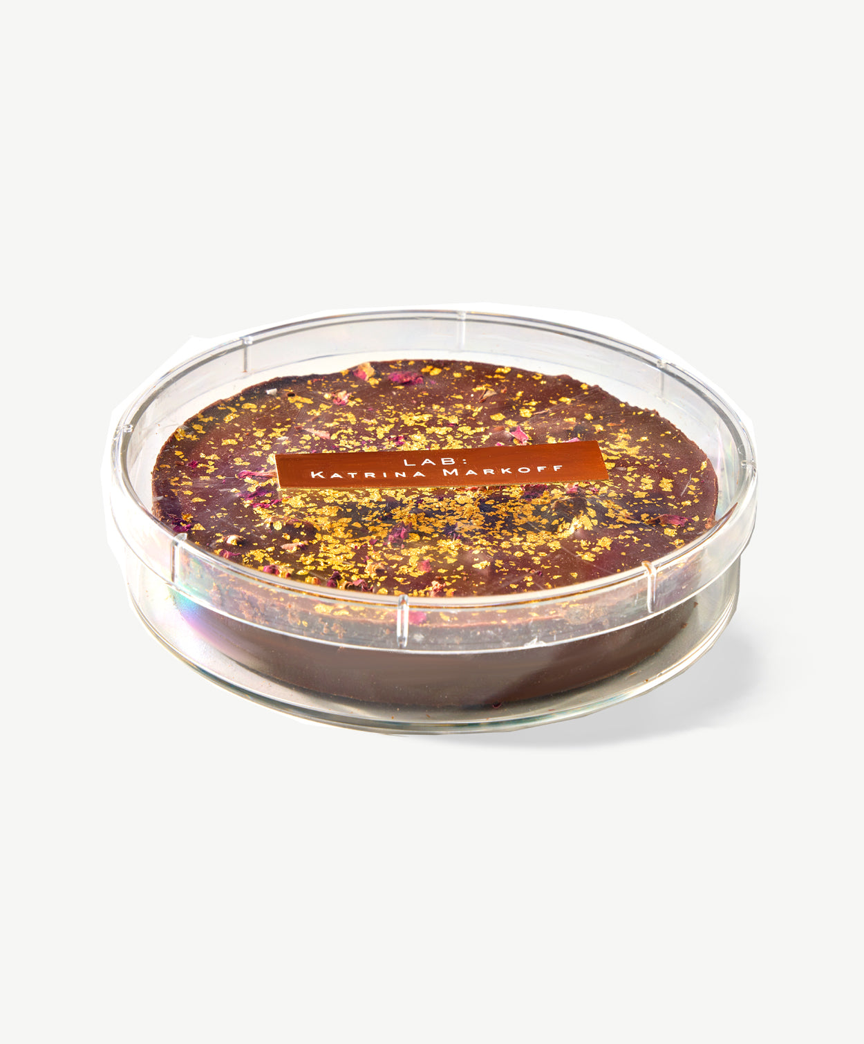 A large chocolate marshmallow disc adorned with gold leaf, pistachios and dried rose petals sit's in a clear case labeled: Lab, Katrina Markoff on a white background. 