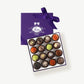 An open purple candy box embossed in silver foil tied with a purple ribbon bow sits open displaying sixteen dark chocolate truffles adorned in brightly colored spices and chopped nuts on a grey background.