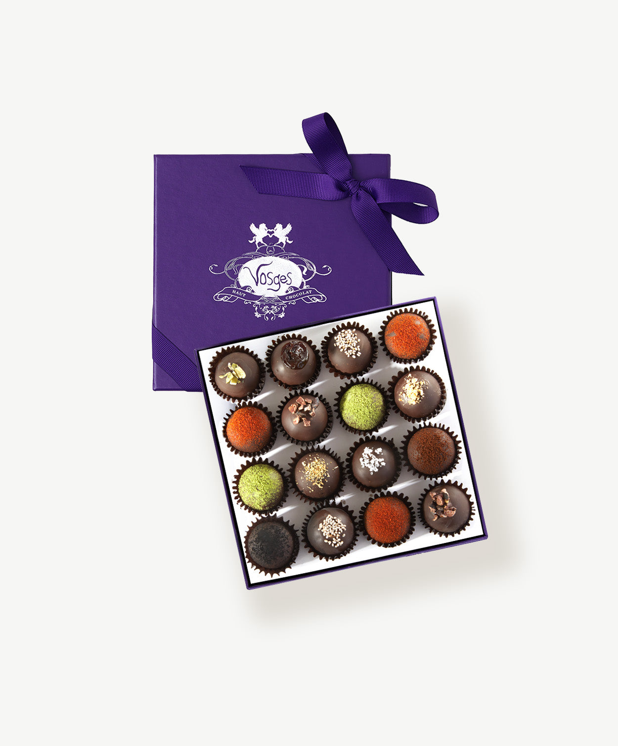 An open purple candy box embossed in silver foil tied with a purple ribbon bow sits open displaying sixteen dark chocolate truffles adorned in brightly colored spices and chopped nuts on a grey background.