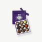 A small purple candy box tied with a purple ribbon bow sit's open displaying three rows of dark chocolate truffles adorned in colorful toppings and chopped nuts on a light grey background.