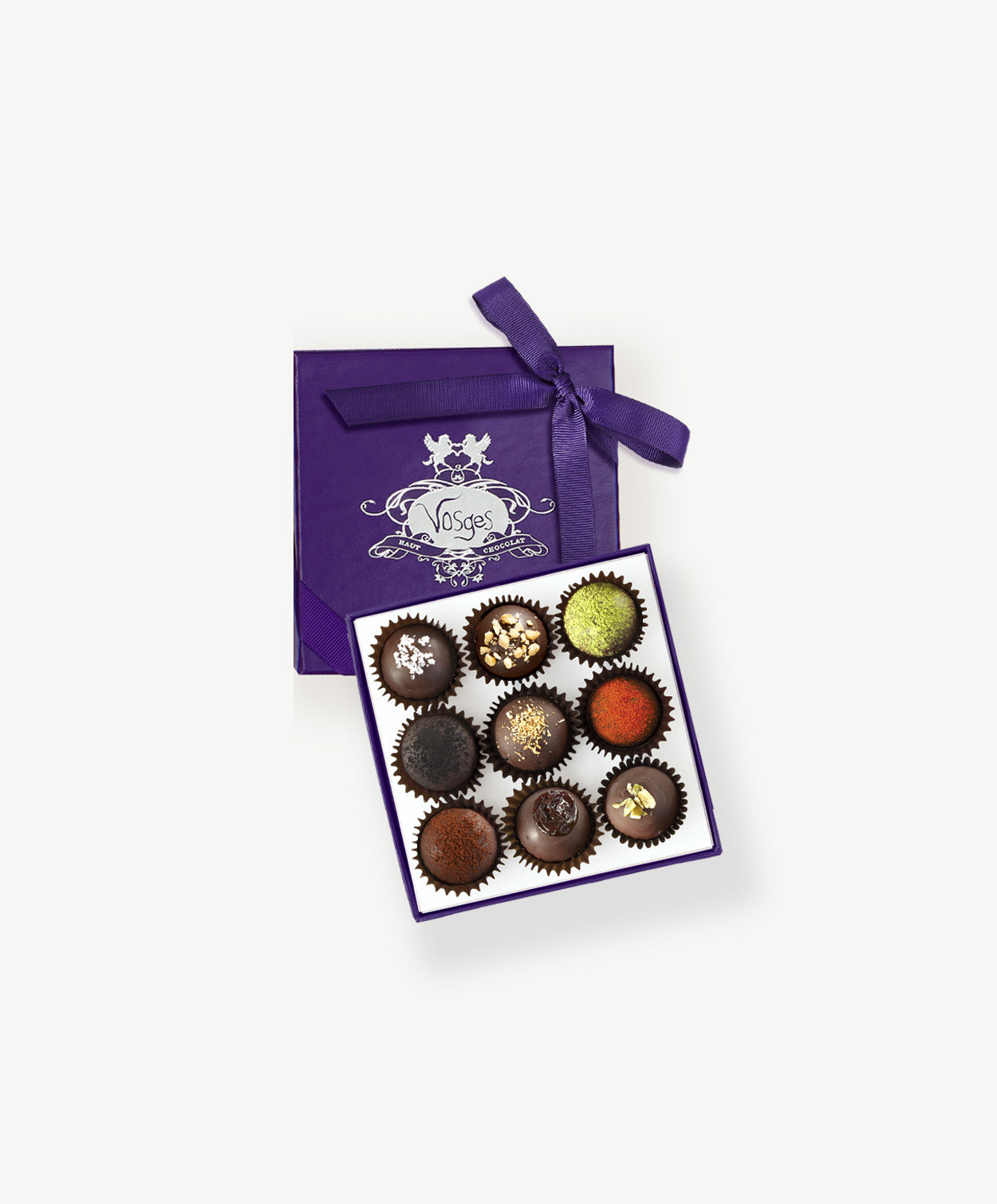 A small purple candy box tied with a purple ribbon bow sit's open displaying three rows of dark chocolate truffles adorned in colorful toppings and chopped nuts on a light grey background.