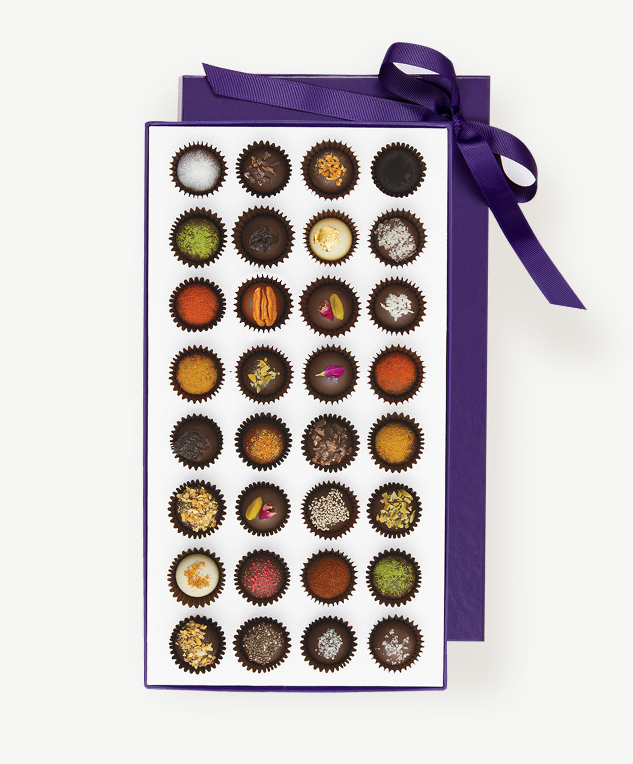 Large purple candy box of Vosges Haut-Chocolat sits open displaying thirty two chocolate truffles adorned with colorful spices, chopped nuts and dried flowers tied with a purple ribbon bow on a grey background.