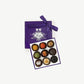 Open purple chocolate box of Vosges haut-chocolat exotic truffles adorned in brightly colored spices and toppings tied with a purple ribbon bow on a white background.