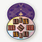 A Round purple box embossed in gold foil sits open displaying eighteen butter caramels enrobed in dark and milk chocolate with colorful nuts and sugar toppings on a grey background.