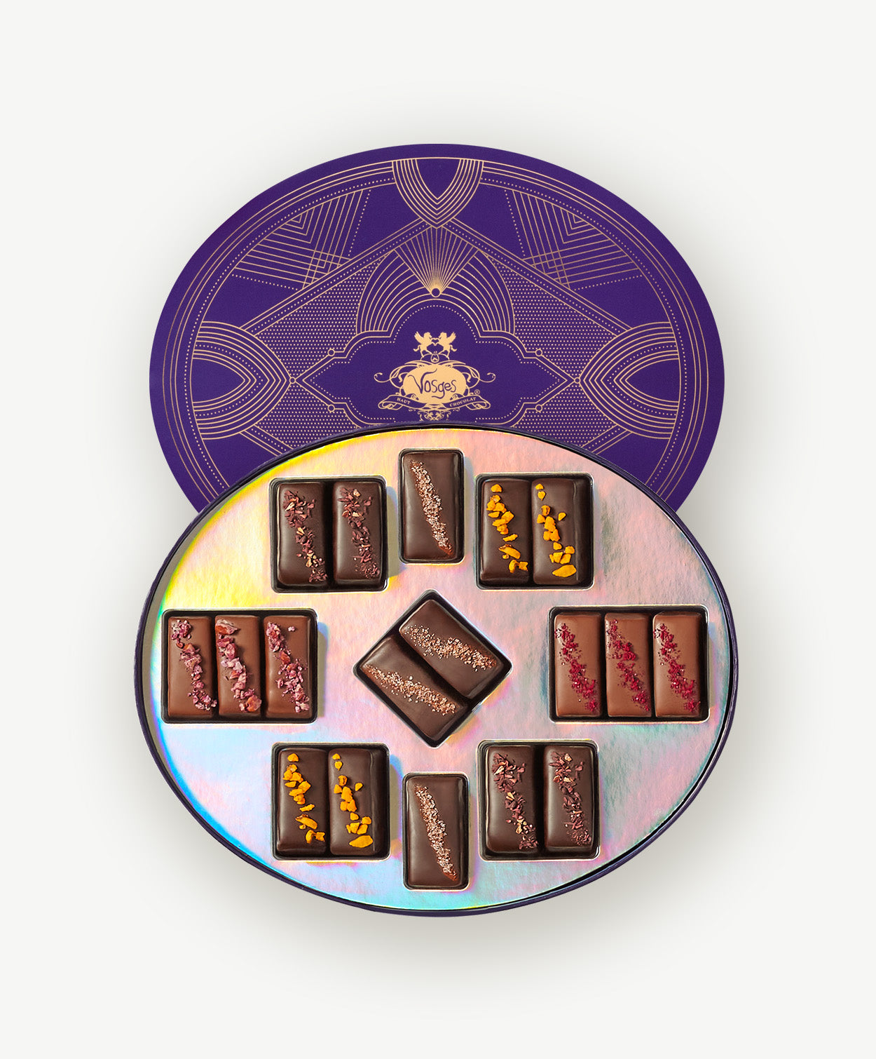 A Round purple box embossed in gold foil sits open displaying eighteen butter caramels enrobed in dark and milk chocolate with colorful nuts and sugar toppings on a grey background.