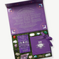 A large, purple giftbox sits open displaying a deck of meditation cards, several crystals, precious stones and  a Vosges Meditation chocolate truffle collection embossed with silver foil tied with a purple ribbon bow nestled in green moss on a grey background.