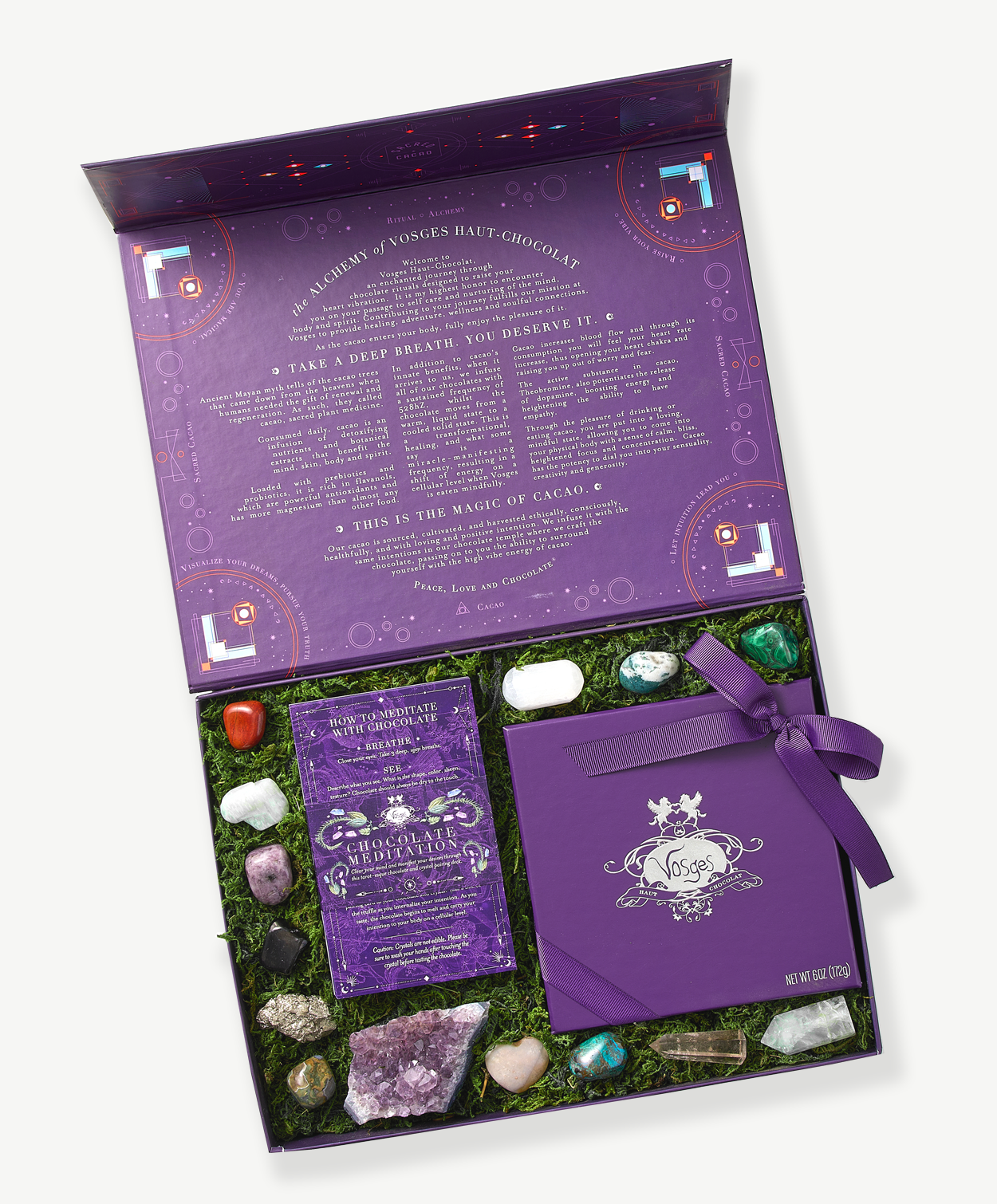 A large, purple giftbox sits open displaying a deck of meditation cards, several crystals, precious stones and  a Vosges Meditation chocolate truffle collection embossed with silver foil tied with a purple ribbon bow nestled in green moss on a grey background.