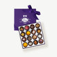 A purple candy box embossed with silver foil and tied with a deep purple ribbon bow sits open displaying sixteen Vosges milk chocolate truffles adorned in brightly colored toppings on a grey background.