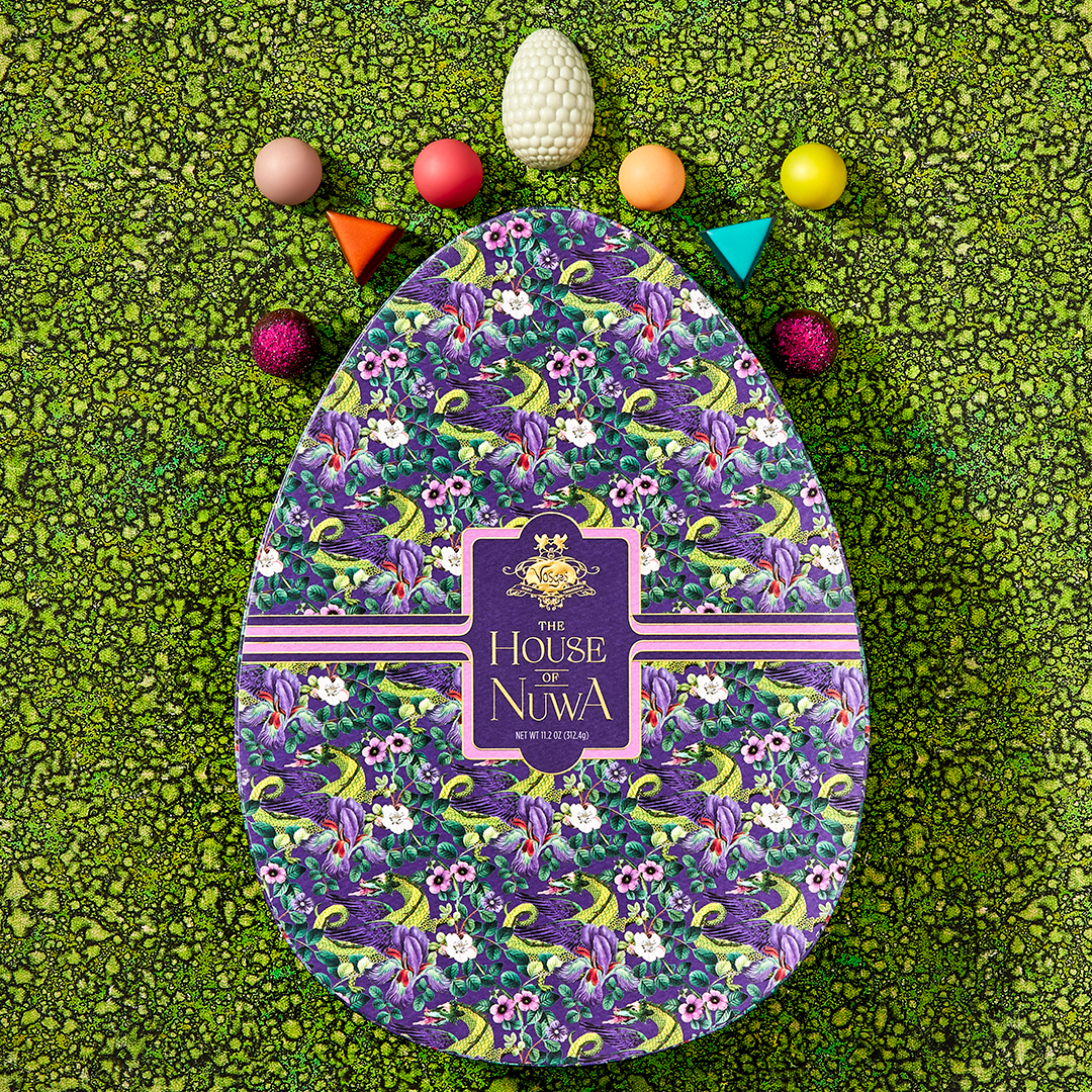 Egg-shaped purple candy box decorated in green dragons and several colorful Vosges chocolate truffles sit on a grass green background.