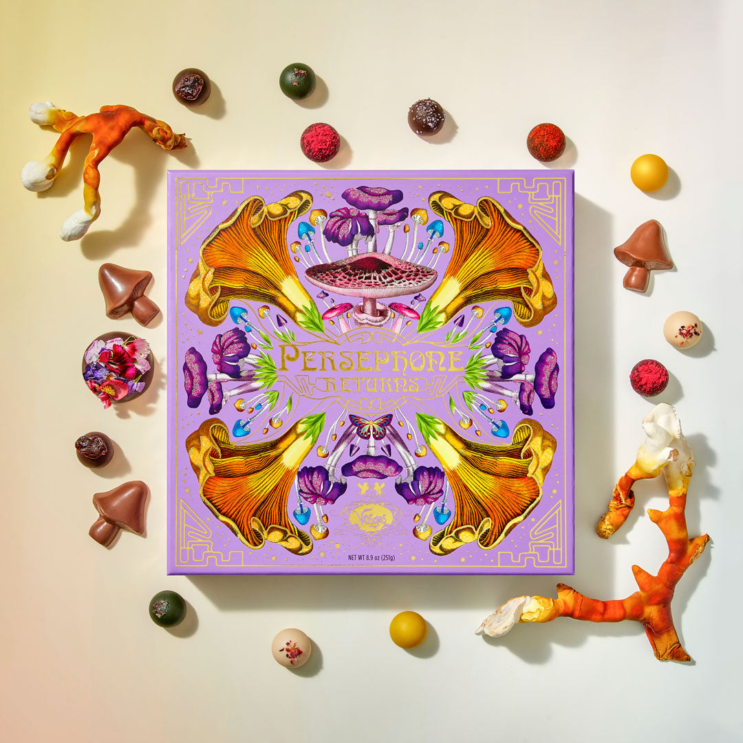 Purple chocolate box brightly decorated in spring flowers reading, "Persephone Returns" in gold foil surrounded by assortment of Vosges Haut-Chocolat Truffles on a beige background. 