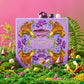Persephone Returns Chocolate Truffle candy box surrounded by white and milk chocolate truffles, sitting in a field of green moss and spring flowers on a pink background.