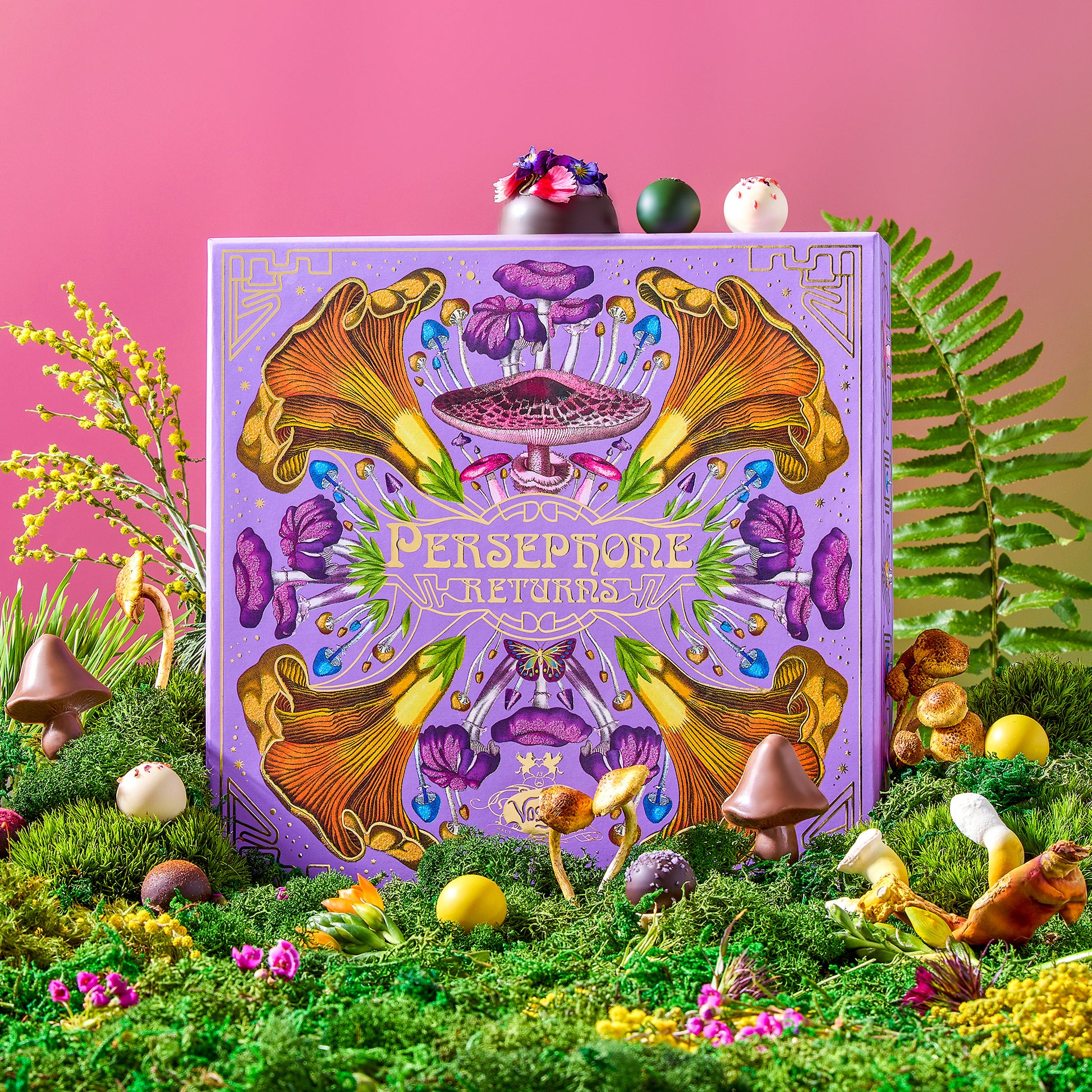 Persephone Returns Chocolate Truffle candy box surrounded by white and milk chocolate truffles, sitting in a field of green moss and spring flowers on a pink background.