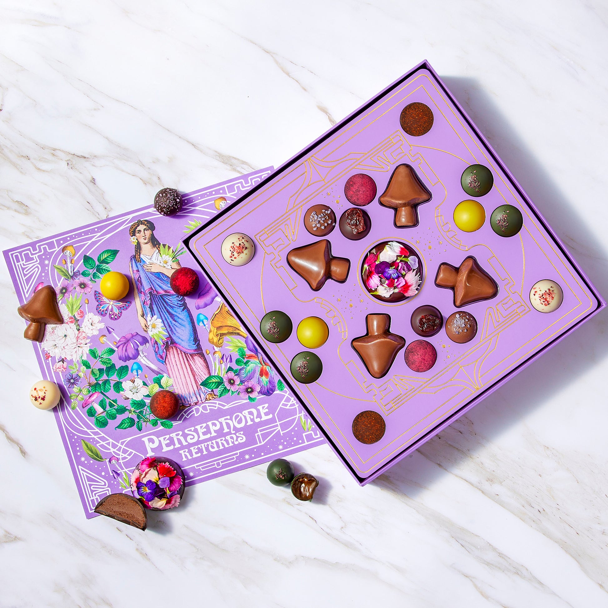 Open box of chocolates and a painting of goddess Persephone surrounded by Vosges Haut-Chocolat Truffles on a grey marble slab.