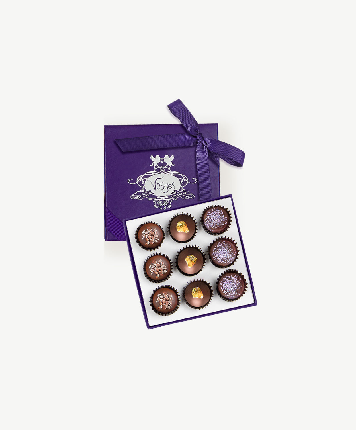 An open purple candy box displaying nine milk chocolate praline truffles topped with gold leaf, candied violets and coco nibs on a white background.