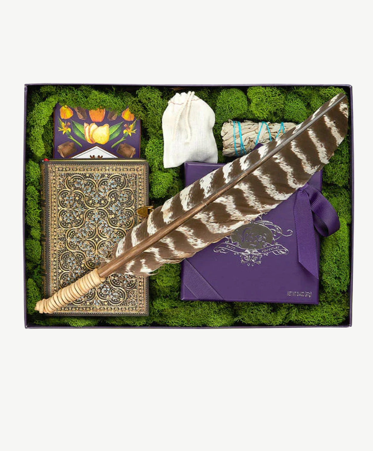 A vosges chocolate truffle collection sits beside an ornately decorated journal, sage bundle, bag of crystals and a stripped turkey feather wrapped with leather cord sit in a moss filled purple gift box on a white background.