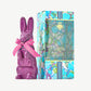 A tall purple chocolate rabbit wearing a pink ribbon bow stands beside a sky blue tie-dyed chocolate box embossed in gold foil on a white background.  