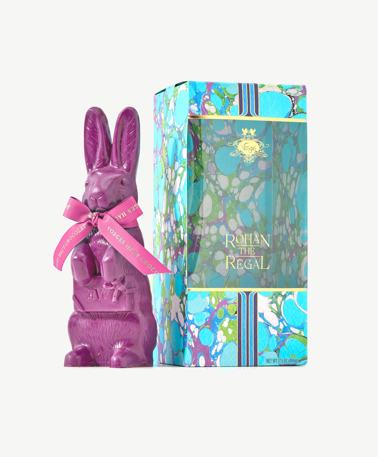 A tall purple chocolate rabbit wearing a pink ribbon bow stands beside a sky blue tie-dyed chocolate box embossed in gold foil on a white background.  