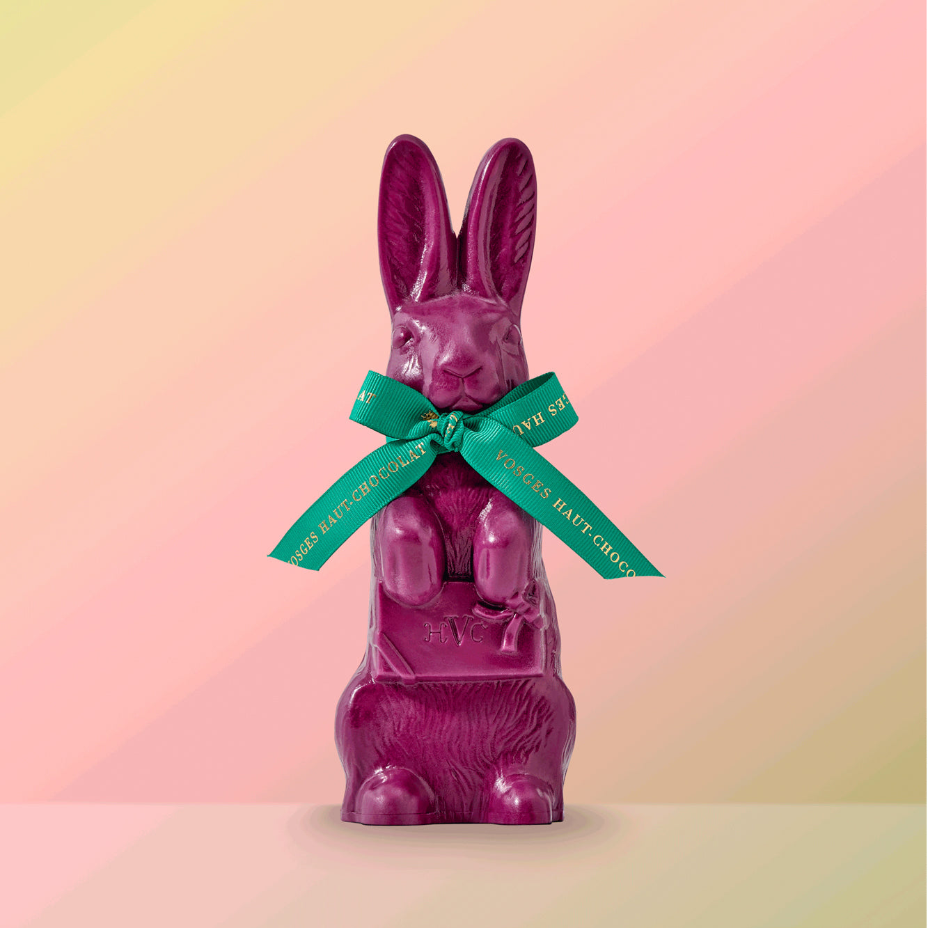A large, purple chocolate rabbit wearing a turquoise and gold ribbon bow stands upright on a soft, pink background.