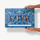 A gigantic two pound crispy peanut butter chocolate bar in a blue tie-dyed wrapper reading, "Love" held in a Caucasian woman's hands.  