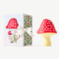 A giant chocolate fly agaric mushroom, red with white polka dots sitting next to a white candy box decorated with red spotted mushrooms, tied with a white ribbon on a white background.