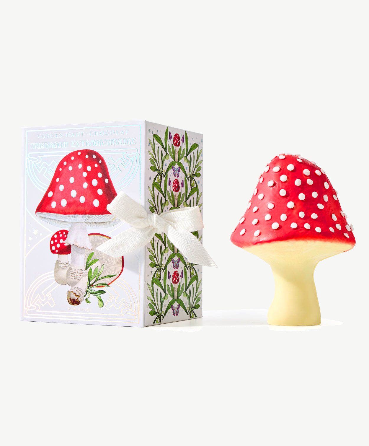 A giant chocolate fly agaric mushroom, red with white polka dots sitting next to a white candy box decorated with red spotted mushrooms, tied with a white ribbon on a white background.