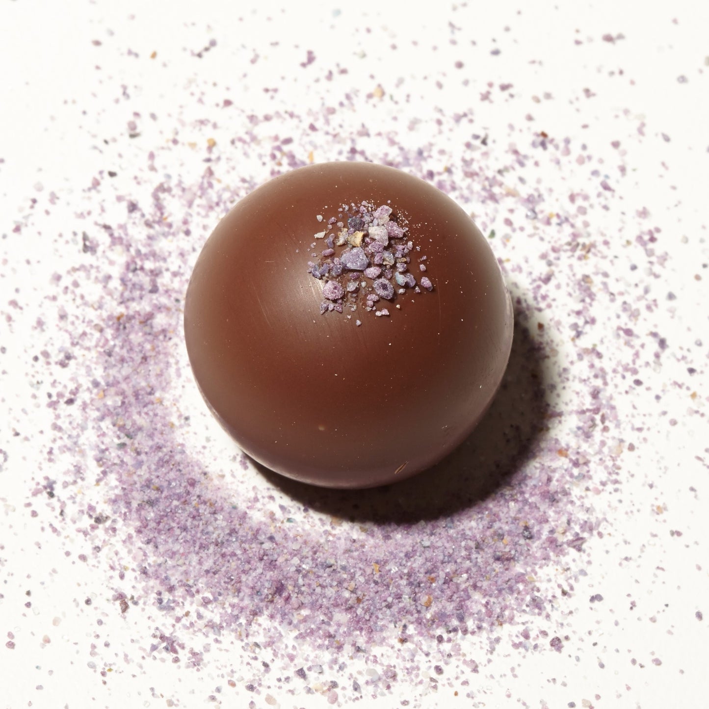 Close-up view of a Vosges Violet truffle coated in milk chocolate and topped with purple candied violet flowers on a white background.