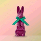 An animated, three hundred sixty degree view of a large purple chocolate rabbit wearing a turquoise and gold ribbon bow spinning in place on a light pink background.