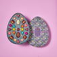 An opened purple and green chocolate box displaying twenty five unique, colorful Vosges truffles on a light purple background. 